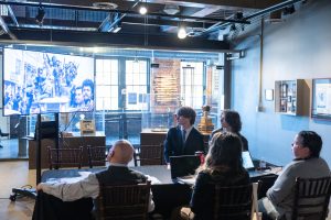 Five people sit at a table in a modern room, viewing a large screen displaying a historic black-and-white group photograph. *AI generated alt text