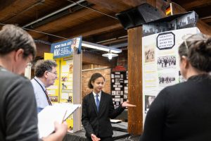 A student in formal clothing presents a German history project to three adults taking notes, with colorful display boards behind them. *AI generated alt text