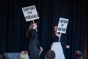 Two women stand on a stage with a black curtain, holding protest signs reading SUPPORT THE MATCHGIRLS and OUR FIGHT IS YOUR FIGHT. *AI generated alt text