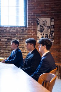 Three young men in suits sit at a wooden table in a brick-walled room with barred window and black-and-white photo behind them. *AI generated alt text