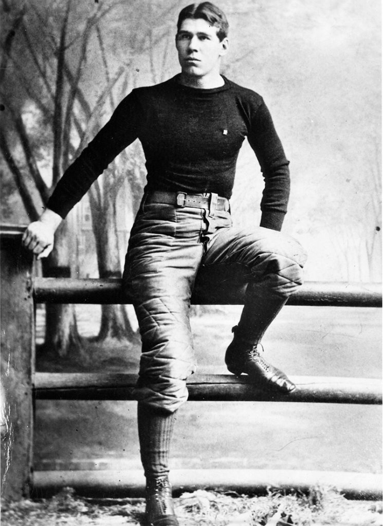 A young man in an early 1900s American football uniform stands with one foot on a wooden fence, trees and misty landscape behind him. *AI generated alt text
