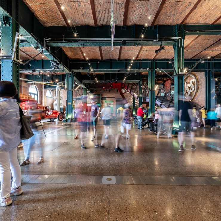 People walk through an industrial-style history museum with exposed brick ceilings, green beams, and lively exhibit displays. *AI generated alt text
