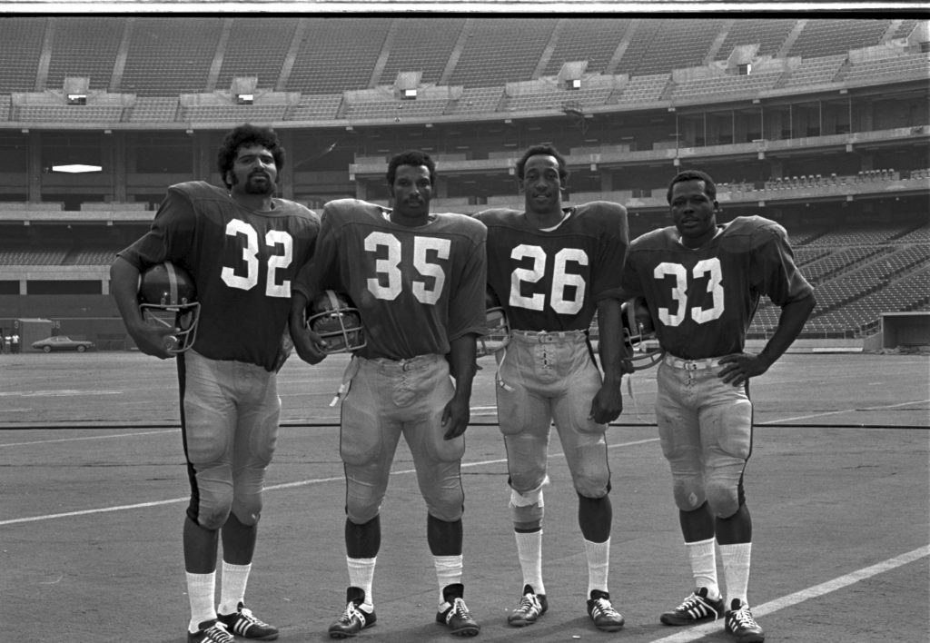 Four American football players in uniform stand on a field in an empty stadium, holding helmets. Jerseys show numbers 32, 35, 26, 33. *AI generated alt text