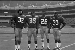Four American football players in uniform stand on a field in an empty stadium, holding helmets. Jerseys show numbers 32, 35, 26, 33. *AI generated alt text