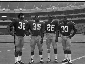Four American football players in uniform stand on a field in an empty stadium, holding helmets. Jerseys show numbers 32, 35, 26, 33. *AI generated alt text
