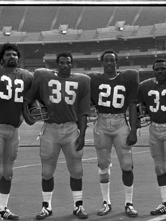 Four American football players in uniform stand on a field in an empty stadium, holding helmets. Jerseys show numbers 32, 35, 26, 33. *AI generated alt text