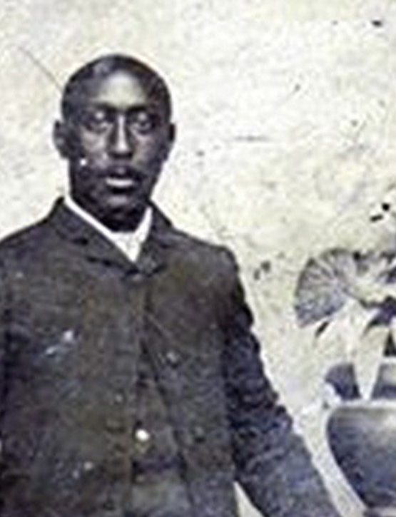 Vintage black-and-white photo of a Black man in a suit standing by a pedestal with a potted plant; aged, faded background. *AI generated alt text