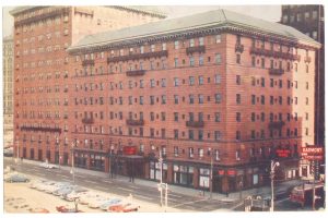 A large brick hotel on a city street, vintage cars parked by the curb, and businesses at street level with a red “HARMONY” neon sign. *AI generated alt text