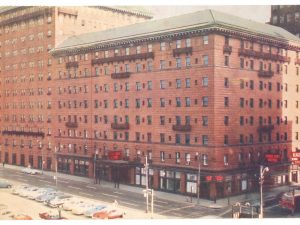 A large brick hotel on a city street, vintage cars parked by the curb, and businesses at street level with a red “HARMONY” neon sign. *AI generated alt text