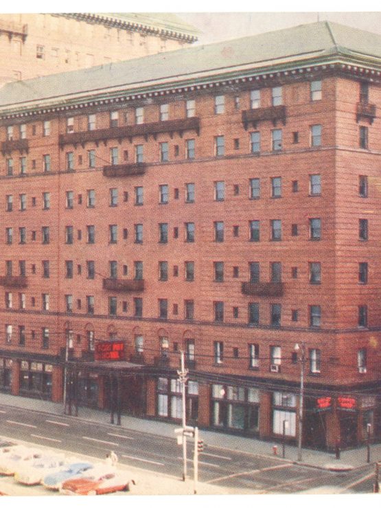 A large brick hotel on a city street, vintage cars parked by the curb, and businesses at street level with a red “HARMONY” neon sign. *AI generated alt text