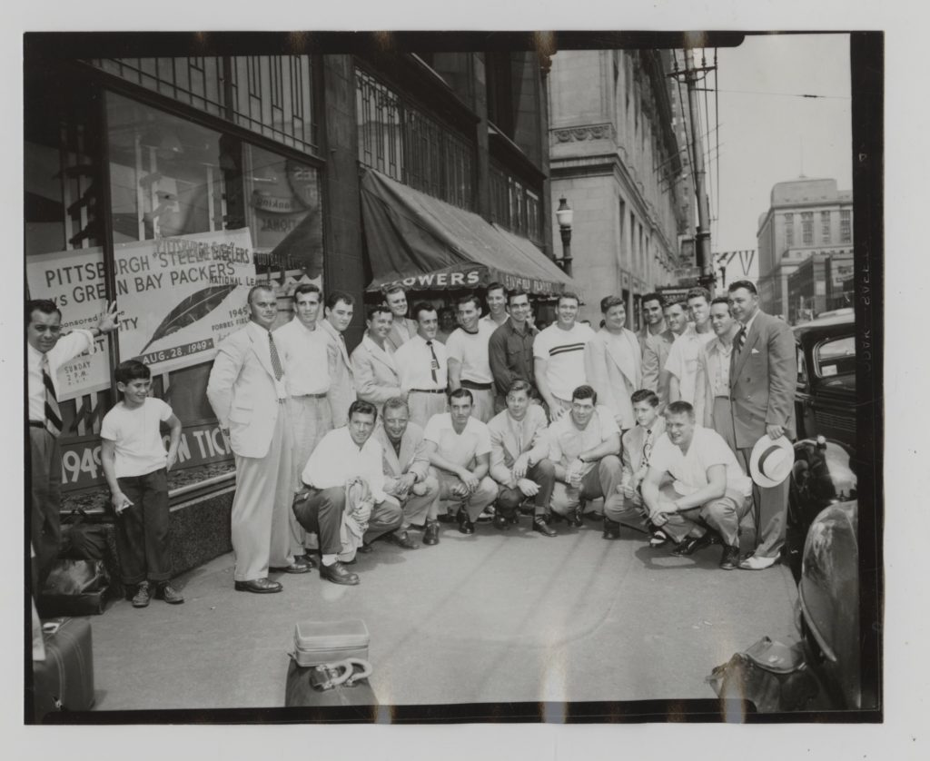 A group of men posing on a city sidewalk in front of a ticket office with a football game sign, buildings and cars behind them. *AI generated alt text