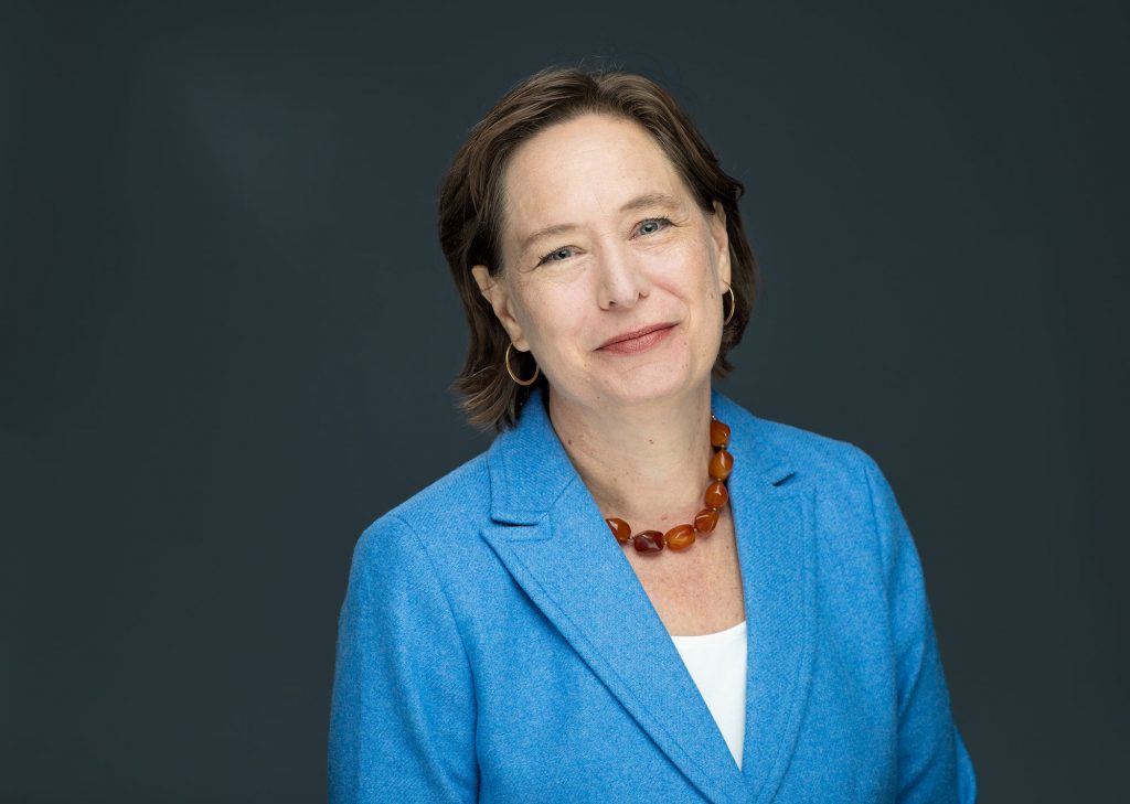 A woman with short brown hair, blue blazer, white top, amber necklace, and hoop earrings poses against a dark gray background. *AI generated alt text