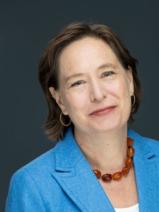A woman with short brown hair, blue blazer, white top, amber necklace, and hoop earrings poses against a dark gray background. *AI generated alt text
