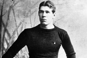 A vintage black-and-white photo of a young man in a dark shirt and belted pants, standing with a serious look before a studio backdrop. *AI generated alt text