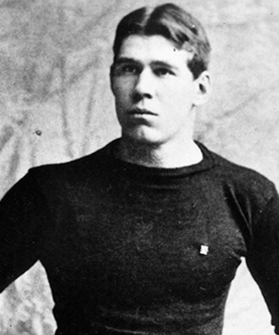 A vintage black-and-white photo of a young man in a dark shirt and belted pants, standing with a serious look before a studio backdrop. *AI generated alt text