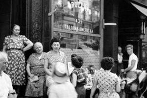 A group of men, women, and children gather outside an Italian American shop; some are seated, others stand or walk, with a guitarist nearby. *AI generated alt text