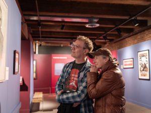 Two visitors stand together in a museum gallery during civic season, viewing framed artwork on blue and red walls with wooden beams. *AI generated alt text