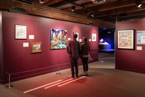 Two people stand in a dimly lit history museum gallery with maroon walls, viewing colorful paintings and framed historical displays. *AI generated alt text