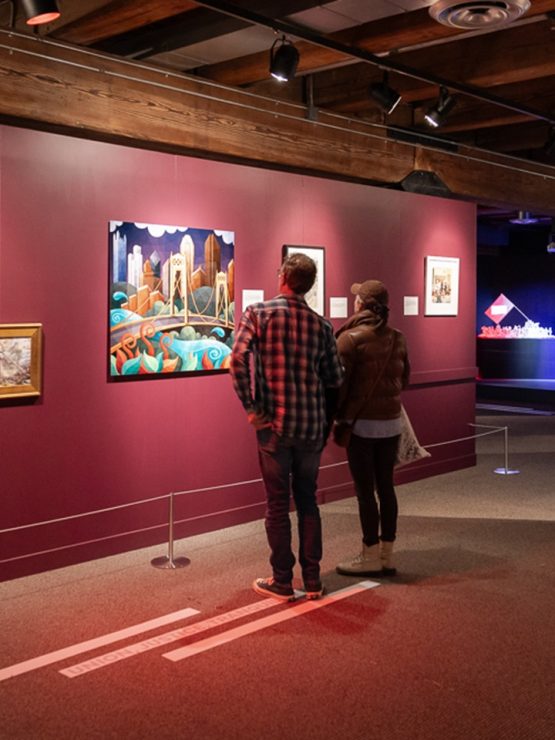 Two people stand in a dimly lit history museum gallery with maroon walls, viewing colorful paintings and framed historical displays. *AI generated alt text