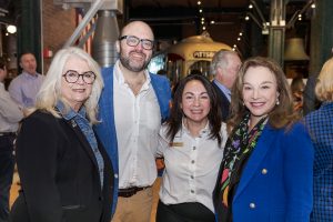 Four adults pose together and smile at an indoor museum event with industrial decor; others and a vintage vehicle are in the background. *AI generated alt text
