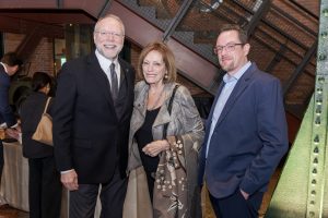 Three adults in formal attire pose at an indoor museum event, flanked by industrial-themed decor in the background. *AI generated alt text