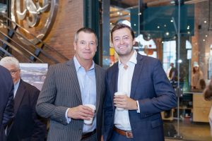 Two men in business suits hold coffee cups and smile indoors, with other people and glass-and-brick office decor in the background. *AI generated alt text