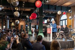 A woman speaks at a podium on stage in an industrial-style museum with balloons, seated audience, classic cars, and three men behind her. *AI generated alt text