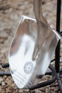 A polished ceremonial shovel with engraved text stands on gravel. The engraving marks a museum expansion groundbreaking in 2026. *AI generated alt text