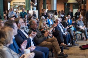 A diverse group gathers in a museum room with exposed brick, watching and applauding an event. A “PITTSBURGH” sign is visible. *AI generated alt text