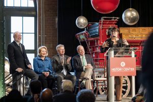 A woman speaks at a podium labeled “History Center” as four people listen and applaud; vintage fire truck and red sphere behind. *AI generated alt text