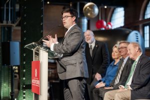 A man in a suit speaks at a podium with a microphone; five people sit on stage behind him. The setting shows industrial decor. *AI generated alt text