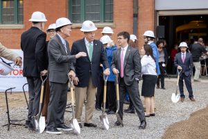 A group in suits and hard hats hold shovels at a groundbreaking ceremony outside a brick museum, with onlookers in the background. *AI generated alt text