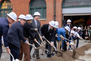 A group in hard hats breaks ground with shovels at a construction site before a brick museum as others watch and take photos. *AI generated alt text