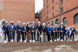 Individuals in business attire and hard hats hold shovels at a construction site, posing for a groundbreaking with brick buildings behind. *AI generated alt text