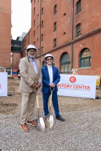 Two men in suits and hard hats holding shovels at a museum groundbreaking ceremony, with brick building and materials visible behind. *AI generated alt text