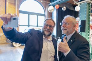 Two men in suits smiling and taking a selfie at a museum event; one gives a thumbs up. Bright background with windows and decorations. *AI generated alt text