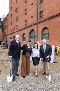 Four people in business attire stand with shovels on a construction site before a brick building; equipment and barriers are behind them. *AI generated alt text