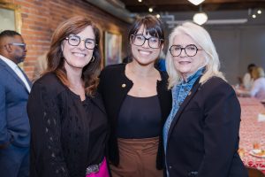 Three women in business-casual attire and glasses pose together at a museum event with exposed brick walls and tables in the background. *AI generated alt text