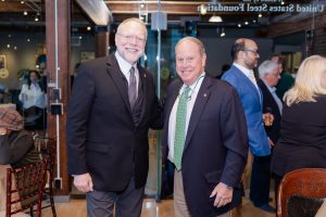 Two men in suits stand together at an indoor museum event, with people mingling and a glass door with text in the background. *AI generated alt text