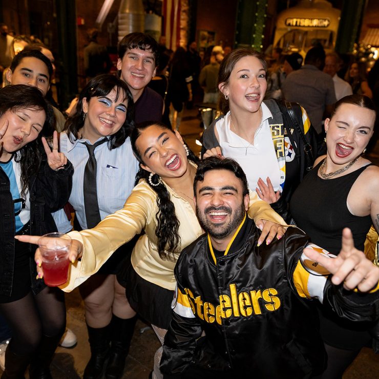 A group of young adults in Pittsburgh Steelers jackets pose at an indoor America 250 event; a crowd and warm lighting are visible. *AI generated alt text