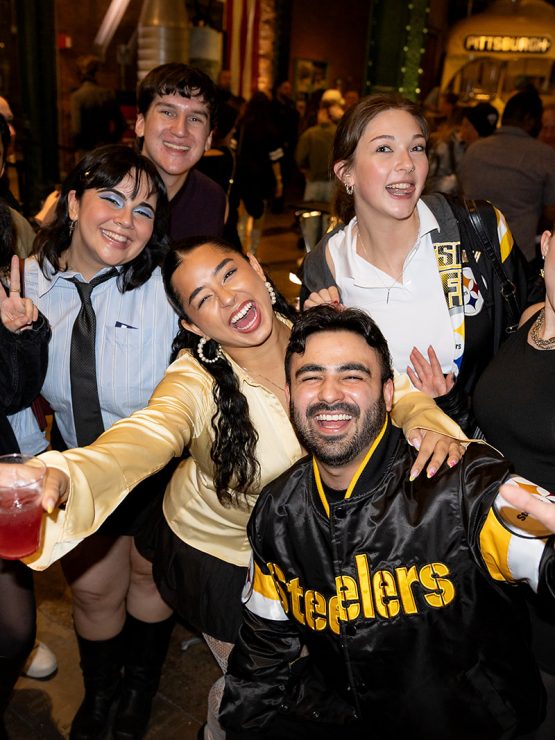 A group of young adults at night, some in Steelers jackets for America 250, pose on a crowded street with one person holding a drink. *AI generated alt text