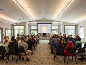 Visitors seated in rows of chairs facing a screen in a bright museum room with large windows, watching a projected presentation. *AI generated alt text