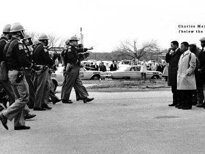 Police officers in riot gear approach a line of Black men standing on a street, with bystanders and parked cars in the background. *AI generated alt text