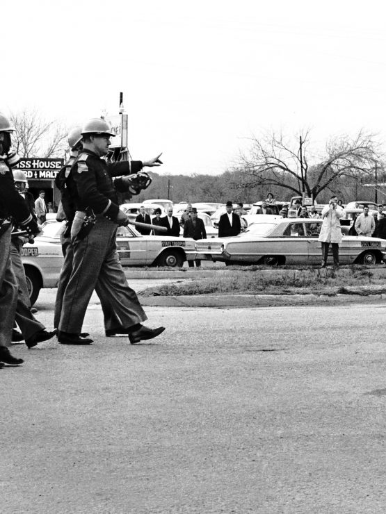Police officers in riot gear approach a line of Black men standing on a street, with bystanders and parked cars in the background. *AI generated alt text