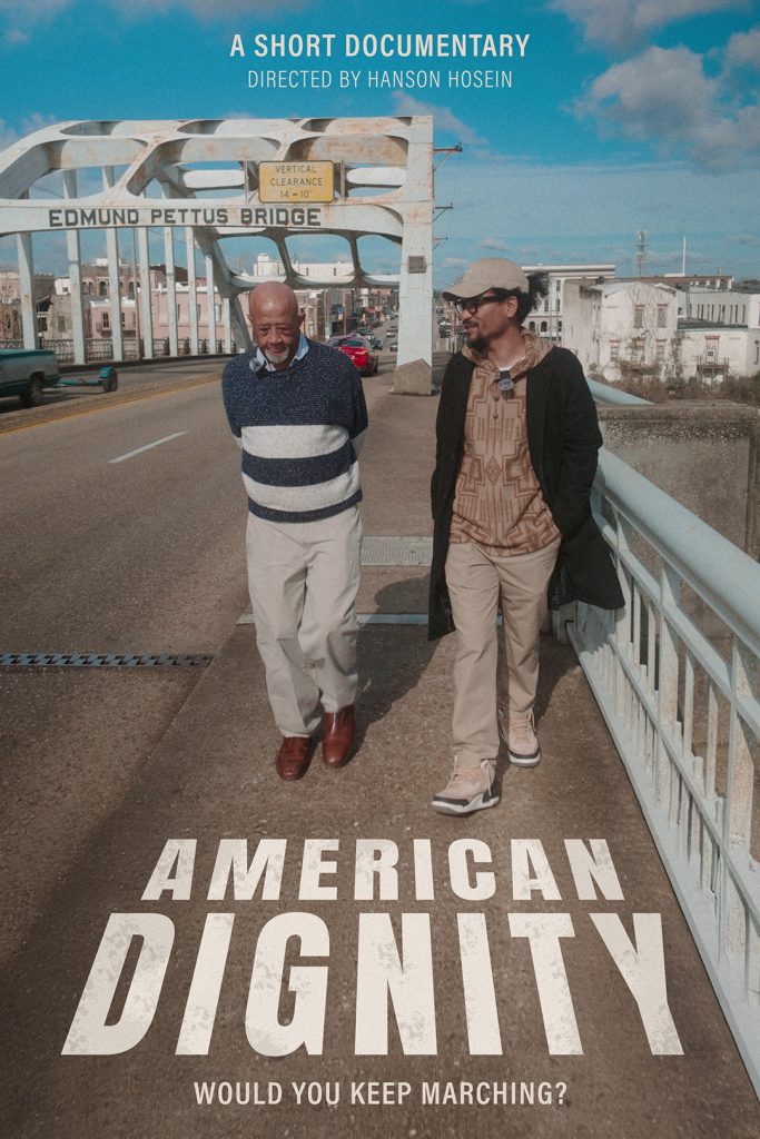 Two men walk and talk on the Edmund Pettus Bridge in Selma, Alabama, under a blue sky. Film title: "American Dignity. *AI generated alt text
