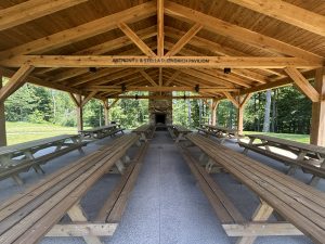 A wooden pavilion with a peaked roof, picnic tables and benches inside, trees behind, and 