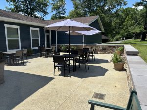 A sunny patio at the museum with black tables, chairs, large umbrellas, potted plants, and a blue house among green trees nearby. *AI generated alt text