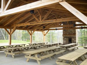 A wooden pavilion with exposed beams, picnic tables, and a stone fireplace in a tree-filled clearing. Sign: “Anthony & Stella D. Church Pavilion.”. *AI generated alt text