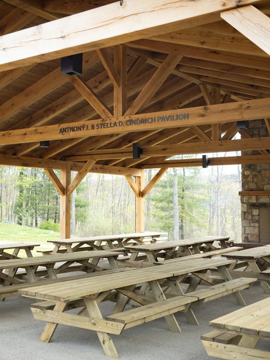 A wooden pavilion with exposed beams, picnic tables, and a stone fireplace in a tree-filled clearing. Sign: “Anthony & Stella D. Church Pavilion.”. *AI generated alt text
