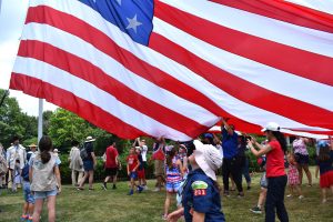 Children and adults at Fort Pitt hold a large American flag overhead during a Civic Season event, with trees and crowds in the background. *AI generated alt text